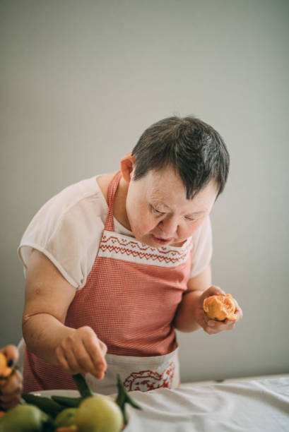 elderly woman with down syndrome is studying in the kitchen peels tangerines stock photo