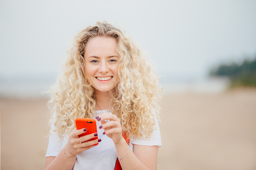 Horizontal shot of good looking young blonde female has curly hair, holds orange smart phone, messages with friends, poses against blurred beach background, rests in tropical country with lover