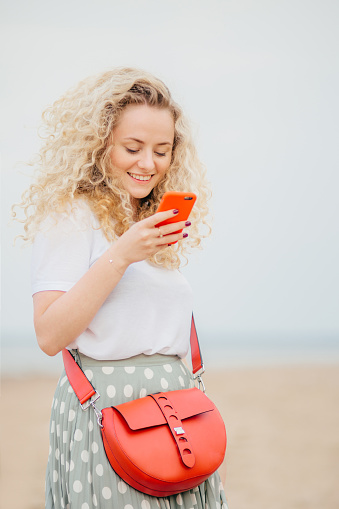 Positive young woman uses modern smart phone, has cheerful expression, makes shopping online, carries bright small bag, has outdoor walk at seaside. People, technology, positiveness concept.