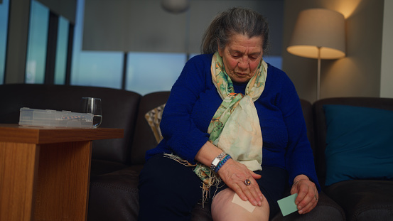 A senior woman is applying a painkiller band on her knee.