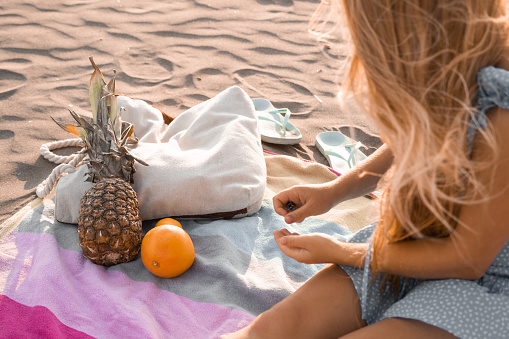 Young blonde woman sitting on a towel at the beach, pineapple, lemon, grapefruit slapper and beach bag in the background.
