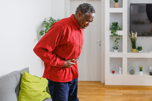 A senior African-American man is leaning and holding his painful stomach, while standing in his living room.