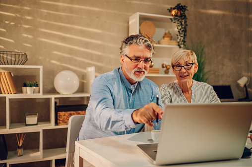 Senior couple sitting at the table at home while online shopping with a laptop and a credit card. Buying via online website and bank card, booking hotel. Focus on a man pointing to a laptop screen.