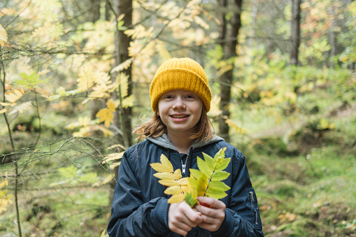 Portrait of a beautiful young girl wearing knitted hat holding yellow leaves in the park. Cute teenage girl having fun on beautiful autumn day outdoors.