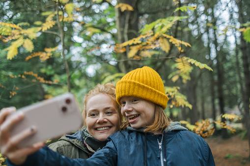 Mother and daughter taking selfie with mobile phone in forest on autumn day. Woman taking selfie with daughter while hiking in a forest.