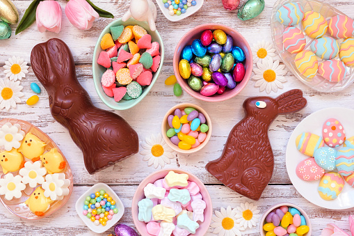 Easter candy table scene. Overhead view over a white wood background. Chocolate bunnies, candy eggs and a variety of sweets.