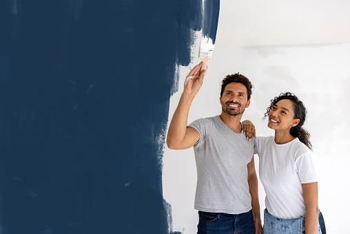 Happy African American couple painting their house with blue paint and using a brush - home improvement concepts