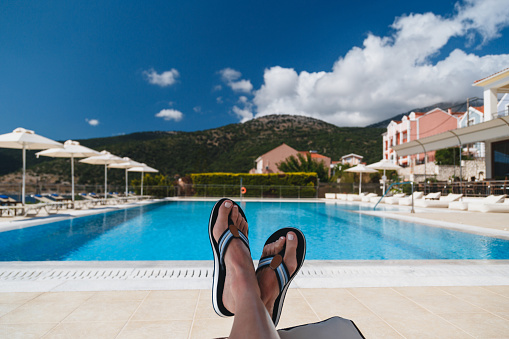 Close up view of young woman feet in blue flip-flops lying on a sun lounger with a swimming pool and blue sky at the background. Kefalonia island, Greece