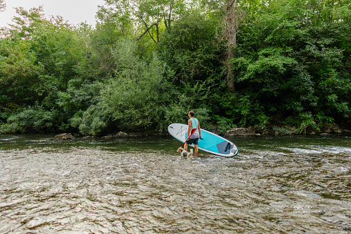 Photo of a young man carrying his standup paddle board down the river and looking for a good spot to paddle, accompanied by his dog