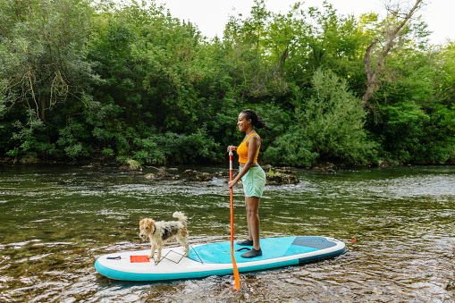 Photo of a young African-American woman riding standup paddle board on the river, while her fox terrier dog is making her company