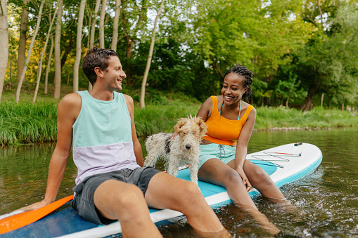 Photo of a young family: a man, a woman and their dog - sitting on a SUP paddle board on a river