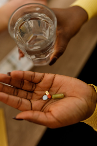 Close up shot of unrecognizable young woman sitting at her desk at the office, holding a glass of water and various pills in the palm of her hand when taking medication.
