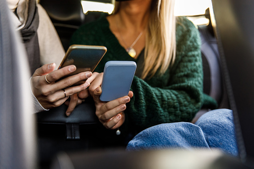 Selective focus shot of two unrecognizable young women, friends, riding in the back seat of a taxi and showing each other memes and data on smart phones,
