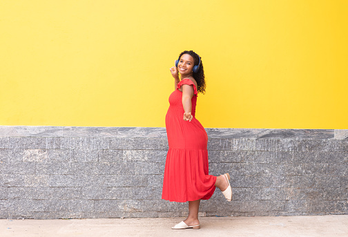 Pregnant black woman walking on the sidewalk and enjoying streaming music on headphones with a yellow wall in the background.