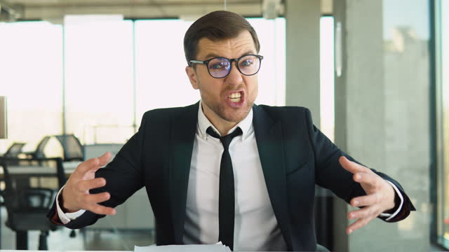 Angry boss shouts at the camera to the employee, dismisses the subordinate, a businessman in a business suit sits at a table in a modern office
