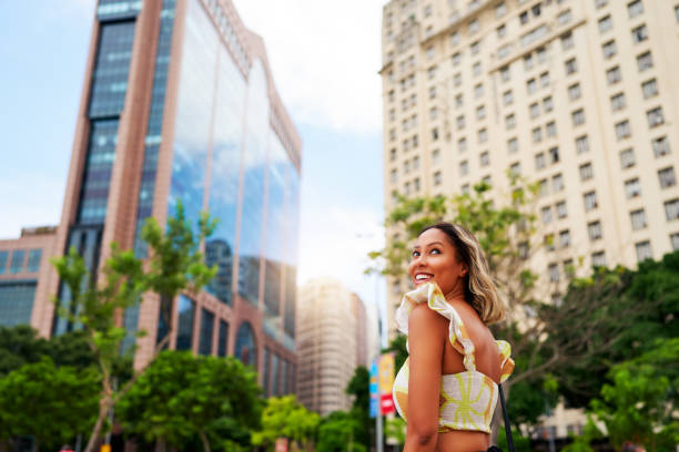 portrait young brazilian black woman smiling looking over her shoulder walking in the city with buildings and trees in the background stock photo