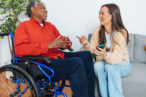A young Caucasian woman holding mobile phone while talking and laughing with a senior African American man in wheelchair