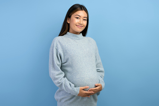 Image of a beautiful young pregnant asian woman posing isolated over blue background. Happy emotions of maternity concept