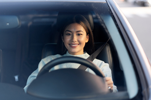 Asian smiling young woman enjoying driving on a beautiful day. Female driver sitting and holding steering wheel with pleasure smile