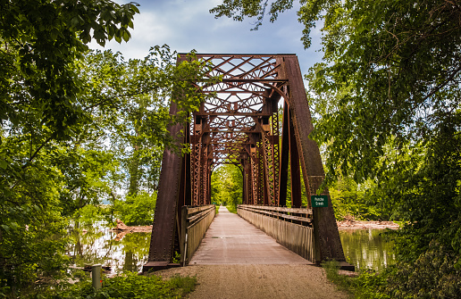 View of old railroad bridge over Midwestern creek turned into recreational trail in summer; trees and rising water on both sides