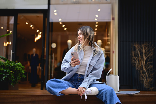 Stylish woman sitting on a wooden beach against a window of a store in a shopping mall.
