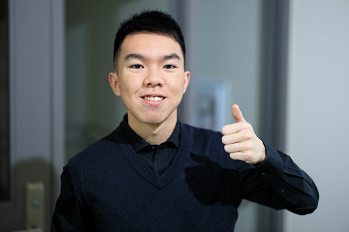 A portrait of a 19-year-old Asian young male university student in a building hallway on campus. He is smiling at the camera giving thumbs up. He is wearing a black shirt and vest sweater. The background is a glass door.