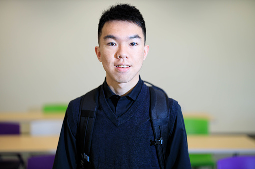 A portrait of a 19-year-old Asian young male university student in a classroom. He is smiling at the camera. He is wearing a black shirt and vest sweater with a backpack. The background is classroom tables and chairs.