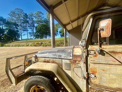 Horizontal still life of rustic + rusted old ute truck parked in farm shed with nature glimpse on rural country property Australia
