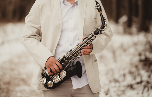 Outdoor Portrait of young male saxophone player. Young Caucasian man with messy hair and moustache, wearing white dress shirt and black coat. Outdoors during the day.