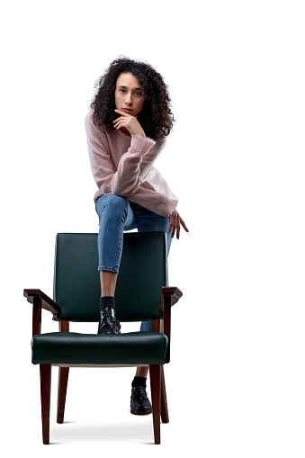 Young curly-haired woman dominates her chair, victorious, with her right leg and left straight and long behind. Leaning on her knee, she touches her chin, staring at the room with an intense expression