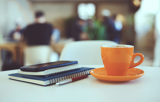 Close-up view, oreange cup of coffee with smartphone, notebook and pen on white table in cafe, freelancer relax with coffee drink after work, blurred  background, vintage collor style