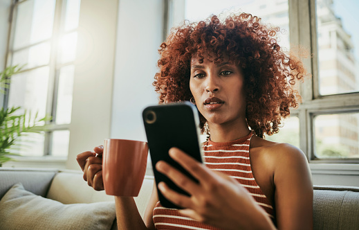 Black woman, coffee cup and reading on sofa with phone for social media, chat app or web in living room. African gen z girl, smartphone ui and texting for networking, email or article on news website