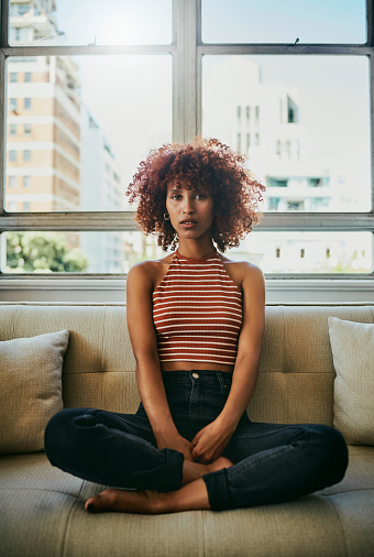 Portrait, relax and sofa with a black woman legs crossed in the living room of her home over the weekend. Window, fashion and apartment with an attractive young female relaxing in her house lounge