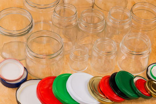 Empty glass jars different sizes with screw thread on mouth and different colored screw lids appropriate diameter for them on a wooden surface