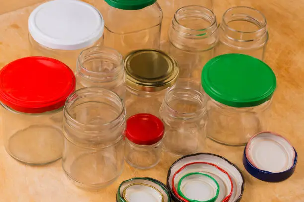 Empty glass jars different sizes with screw thread on mouth and different colored screw lids appropriate diameter for them on a wooden surface