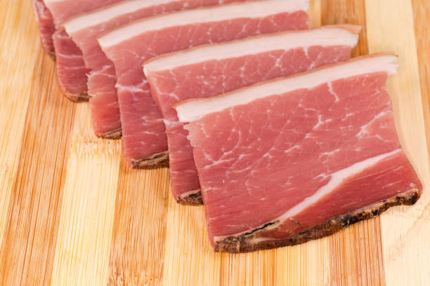 Slices of the dry-cured pork belly on a skin on the bamboo wooden cutting board, fragment close-up in selective focus