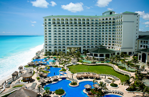 Aerial view of a Cancun resort.  There are several large pool areas and a multi-level hotel at this resort.  An ocean and sandy beach can be seen to the left of the hotel.