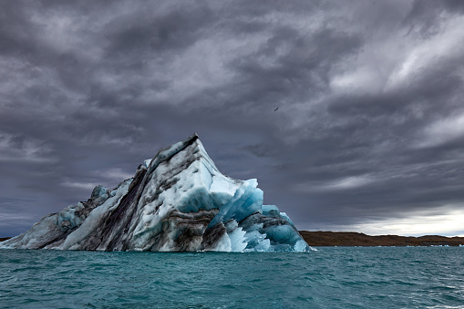 Moody photo of big pointy iceberg. The blue-black iceberg shows layers of old dust and/or volcanic ash fallen on the 