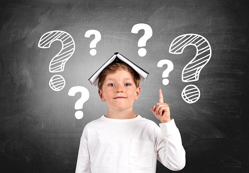 Smiling school boy with a book on head and index finger pointing up, portrait with question marks drawn on chalkboard. Concept of idea and plan