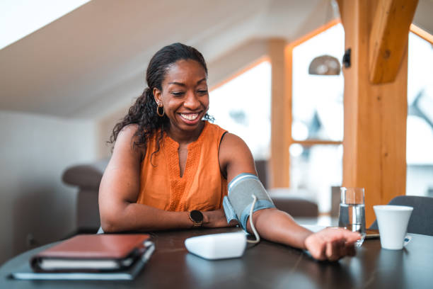 Black Female In 40s Using A Medical Device For Measuring Blood Pressure Mid adult black female using a medical device in a domestic dining room. Measuring blood pressure at home. blood pressure medication stock pictures, royalty-free photos & images