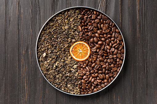 Green dry tea, roasted coffee beans and a slice of dry orange in a round box on a dark wooden background top view. Conceptual composition of green tea and coffee beans with space for text.