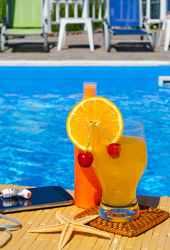 Orange juice with cherries. Mobile phone, headphones and a starfish on the background of the pool. Summer family vacation concept.