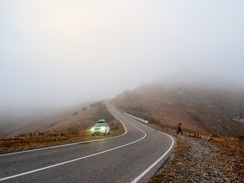Russia. Ingushetia. October 2022.  White car SUV with fog lights turned on is parked next to a scenic route road in a foggy autumn landscape. Luggage trunk box on car roof rack. Sun is in the fog.