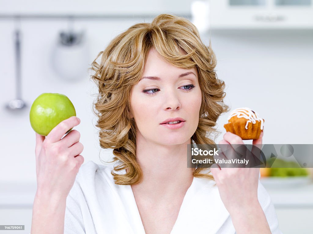 Woman choose between cake and apple Young beautiful woman choose between sweet cake and green apple - in the kitchen 20-29 Years Stock Photo