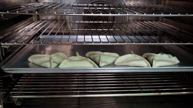 A baker's hand inserts a tray of raw empanadas into an industrial oven.