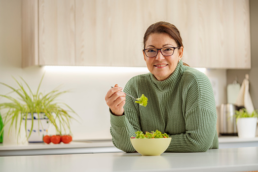 Portrait of a confident mature woman looking at camera and eating healthy  fresh salad. She is standing in domestic kitchen. Selective focus on foreground. High resolution 42Mp studio digital capture taken with Sony A7rII and Sony FE 90mm f2.8 macro G OSS lens.