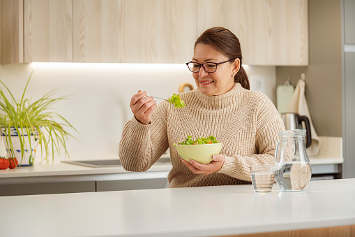 Healthy eating habits. Portrait of a confident mature Hispanic woman eating healthy fresh salad and drinking water. She is sitting in domestic kitchen. Selective focus on foreground. High resolution 42Mp studio digital capture taken with Sony A7rII and Sony FE 90mm f2.8 macro G OSS lens.