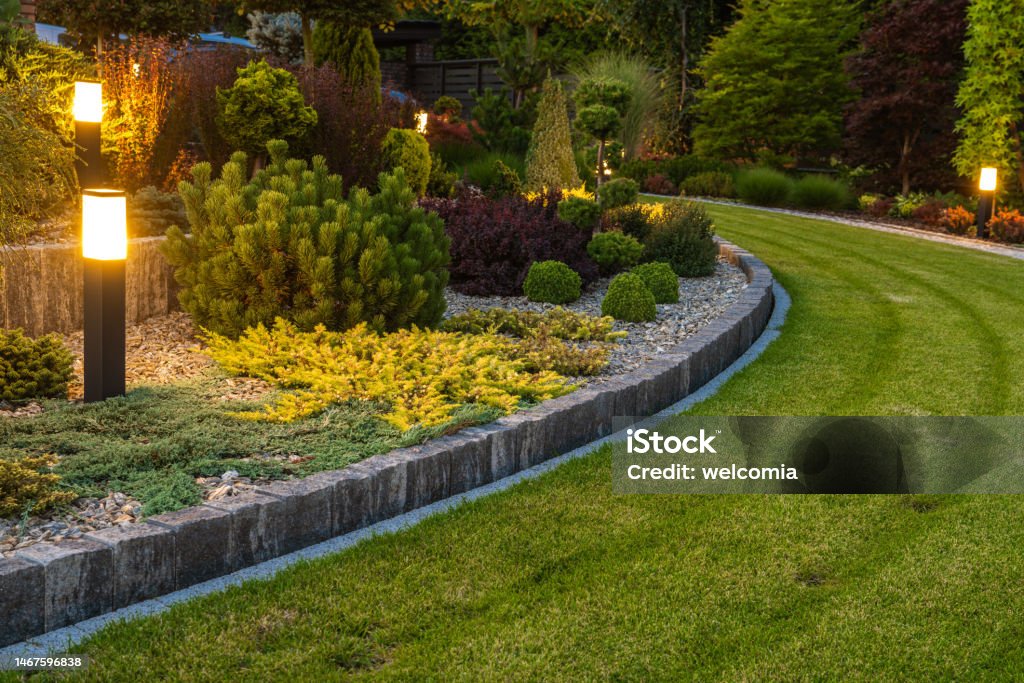 Residential Garden Landscaping Design Idea Professionally Landscaped Backyard Garden with Evenly Mowed Lawn and Trimmed Shrubs Illuminated with Outdoor Bollard Lamps. Landscaped Stock Photo