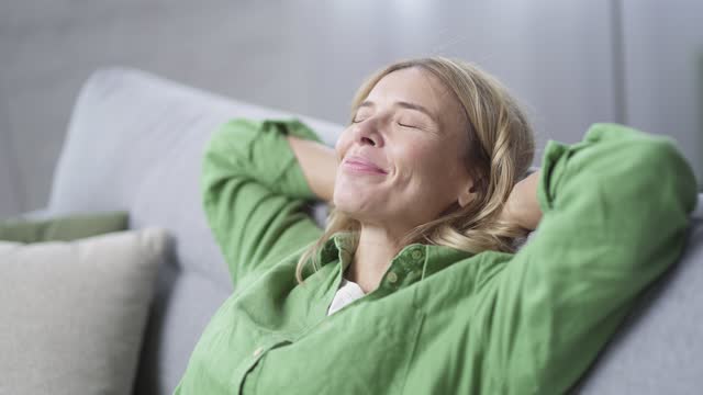 Close up of happy mature blonde woman relaxing sitting on sofa in living room at home. Smiling female in casual clothes stretches with hands behind her head and eyes closed indoors