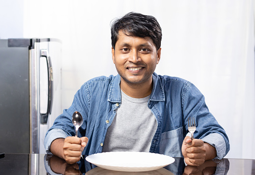 An Indian male sitting beside dining table with empty plate on white background
